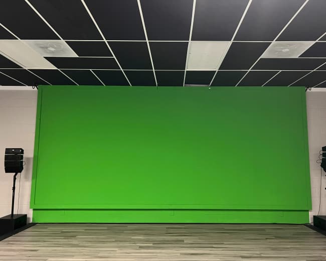 A green backdrop on a wall in a room with black and white ceiling tiles and speakers on either side.