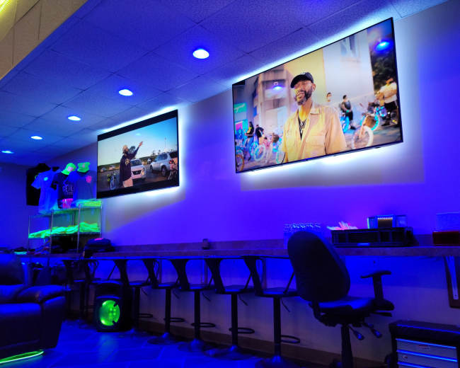 The image shows a brightly lit lounge area with two televisions mounted on the wall, displaying videos, and a bar-like seating arrangement with high stools.