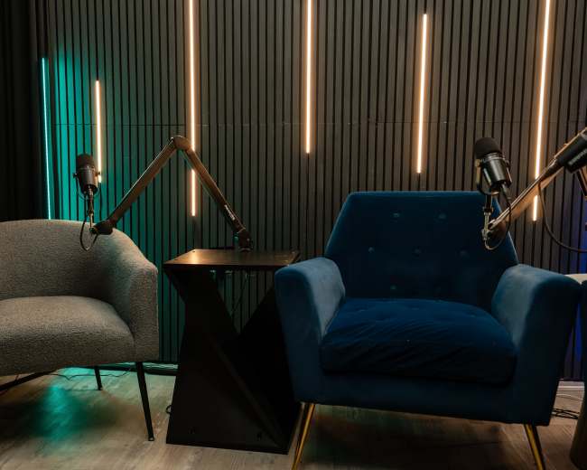 The image shows a podcast studio setup with two chairs, a small black table, and microphones mounted on adjustable arms against a backdrop of vertical wooden panels and illuminated strips.