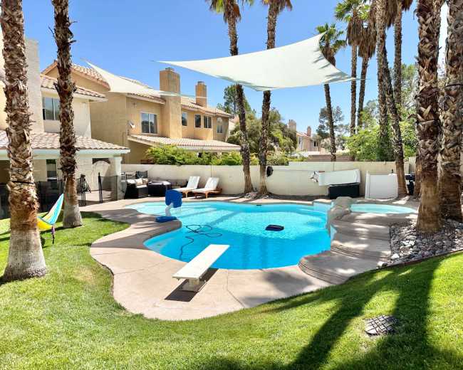 A swimming pool surrounded by palm trees and lounge chairs, with a shade sail above and a grassy area nearby.