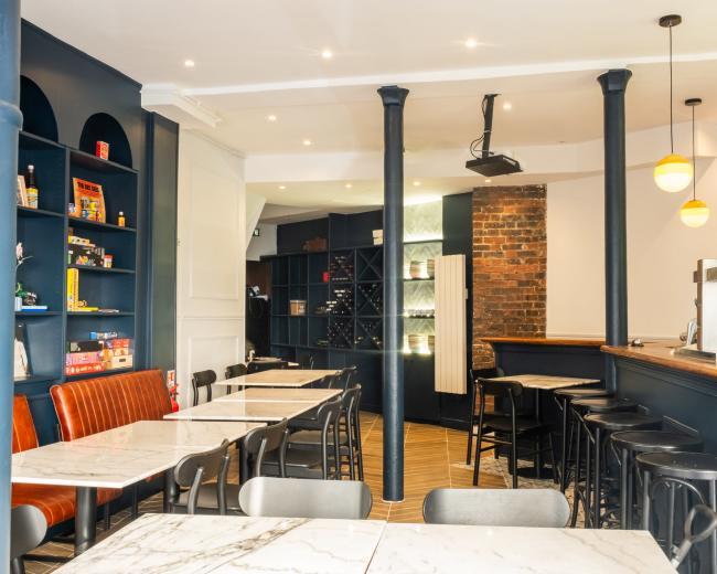 The interior of a café features dark blue walls, wooden accents, and a mix of seating arrangements, including marble-topped tables and high stools at the bar.