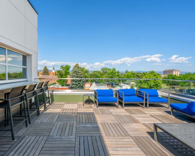 The image shows a rooftop patio with blue lounge chairs and a bar seating area overlooking a green landscape under a clear blue sky.