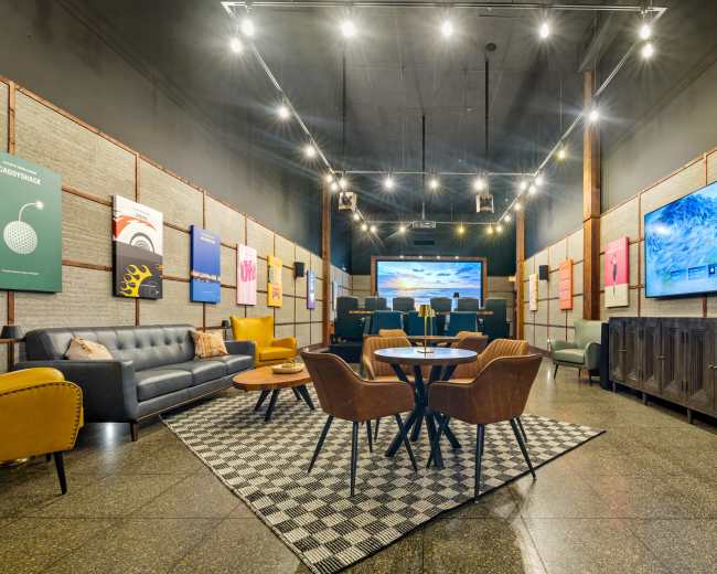 The image depicts a stylish lounge with a cozy seating area featuring a gray sofa, multiple armchairs, a round table, and large screens displaying artwork.