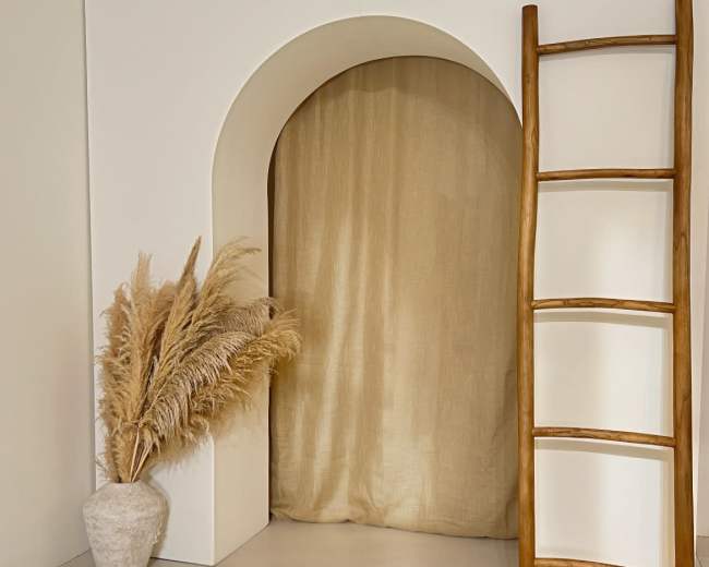 A wooden ladder leans against a white wall beside a potted arrangement of dried pampas grass in a textured vase, with a beige curtain hanging in an arched alcove.