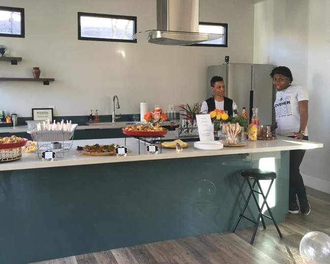 A modern kitchen features a large island with food platters and drinks, while two people stand nearby.
