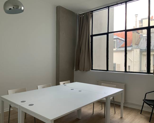 A bright room features a large white table surrounded by chairs, with a window revealing a view of rooftops and a gray wall.
