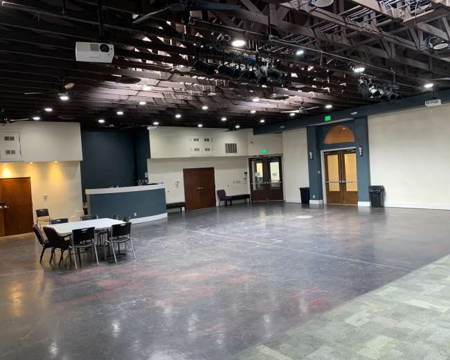The image shows a large, empty multipurpose room with a dark floor, overhead lighting, and a small stage area, featuring tables and chairs arranged on one side.
