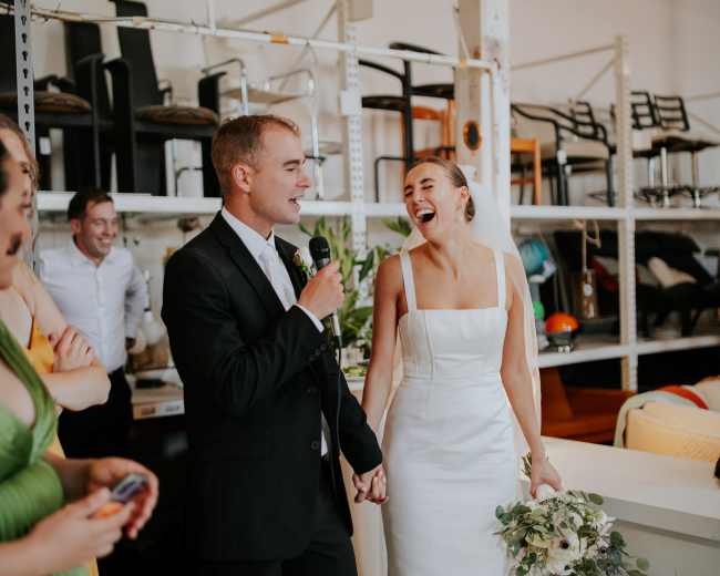 A man in a suit speaks into a microphone while holding hands with a smiling woman in a white wedding dress, surrounded by guests in a bright, industrial-style venue.