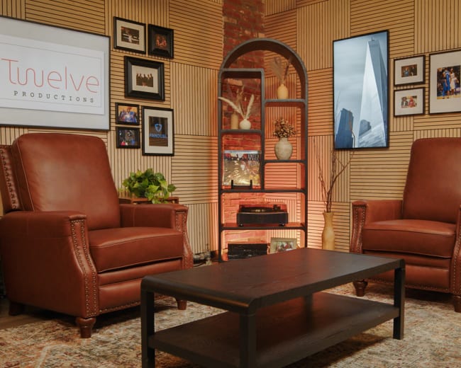 The image shows a stylish interior featuring two brown leather armchairs facing a wooden coffee table, with framed photos and screens mounted on the walls amid wooden paneling.