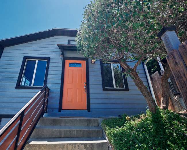 A light blue house features an orange front door and a sloped entrance with wooden steps.