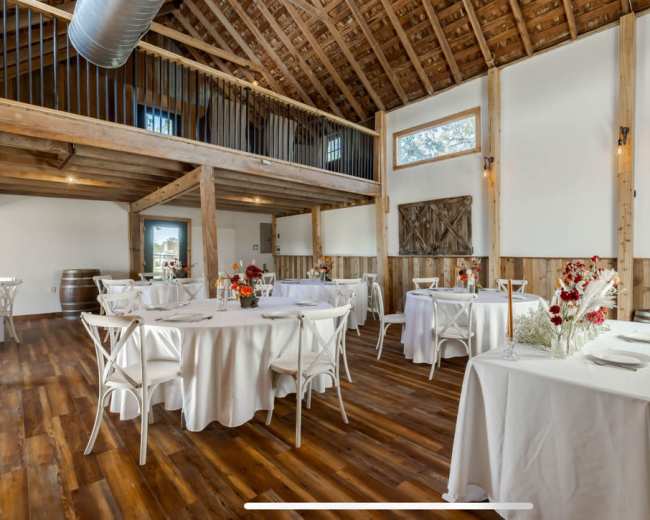 An event space with several round tables covered in white tablecloths, surrounded by wooden beams and walls, with large windows letting in natural light.