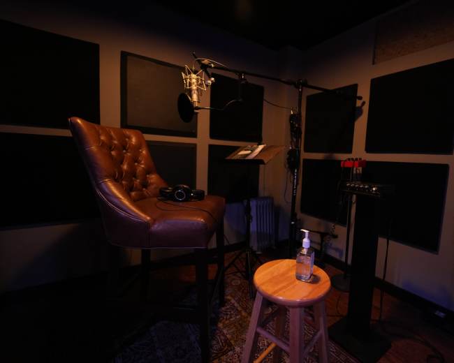The image shows a sound recording studio with a leather chair, a microphone setup, and a wooden stool beside a bottle of hand sanitizer.