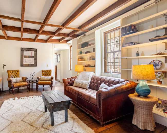 A cozy living room features a brown leather chesterfield sofa, two upholstered armchairs, a wooden coffee table, and shelves filled with decorative items.