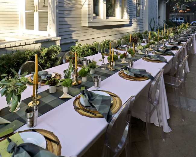 A long outdoor dining table is set with plates, glasses, and candles, aligned next to a house and decorated with greenery and lanterns overhead.