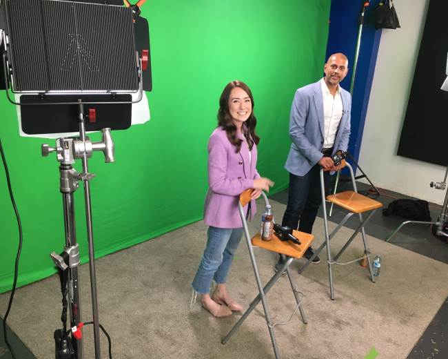 Two individuals stand on a green screen set, one in a purple blazer and the other in a light blue jacket, with chairs and equipment visible around them.