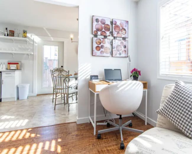 The image shows a small, bright home office with a desk, a chair, and a decorative wall display, adjacent to a dining area.