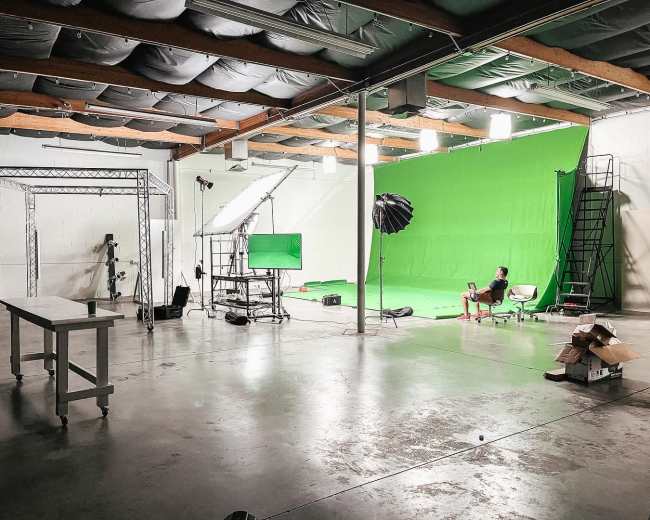 An empty studio space features a green screen setup, lighting equipment, and a person sitting on a chair.