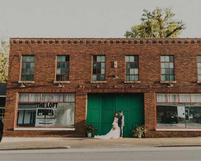 A couple in wedding attire stands in front of a brick building with green doors and large windows.