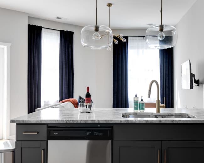 A modern kitchen features a marble countertop, dark cabinetry, and pendant lights above the sink, with bottles and a wine glass on the surface.
