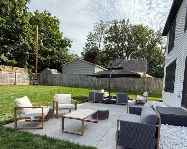 A large outdoor seating area features modern furniture arranged on a stone patio surrounded by grass and trees.
