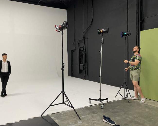 A man in a black suit stands in the center of a photography studio while another man adjusts lighting equipment nearby.