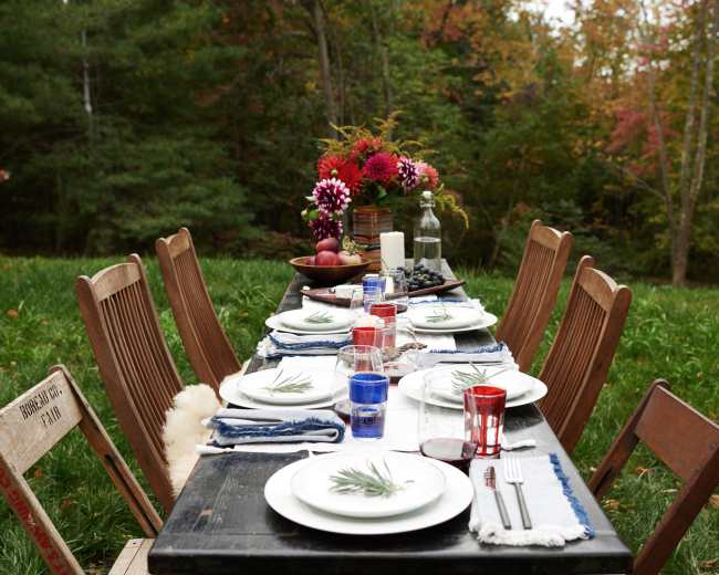 A long outdoor dining table is set with plates, glasses, and a vase of flowers, surrounded by greenery and autumn foliage.