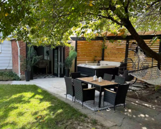 The image shows a backyard with a patio area featuring a table and chairs, surrounded by greenery and a hot tub in a fenced space.