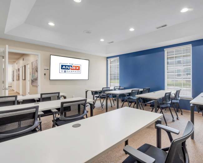The image shows a modern meeting room with multiple tables and chairs arranged for a workshop, featuring a large display screen on the wall.