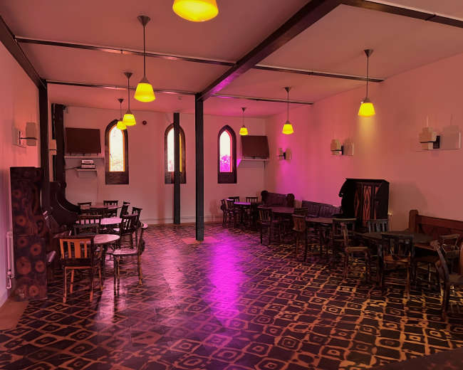 The image shows an empty interior of a café or restaurant featuring wooden tables and chairs, with pink walls and unique patterned flooring illuminated by warm light fixtures.