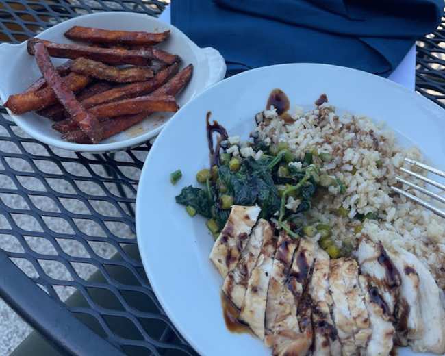 A plate with grilled chicken, rice, sautéed greens, and a side of sweet potato fries on a metal table.