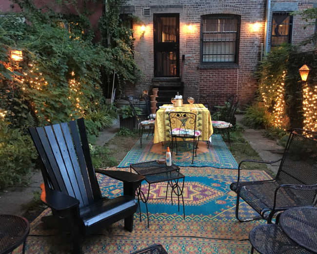 A cozy garden area with a table set for dinner, surrounded by lights and greenery, and featuring various seating options.