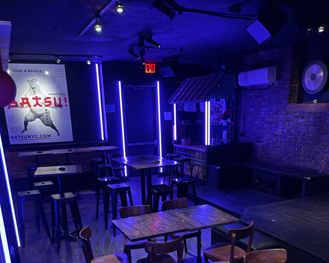The image shows an empty music venue with wooden tables and chairs arranged in a dimly lit space featuring purple neon lighting.