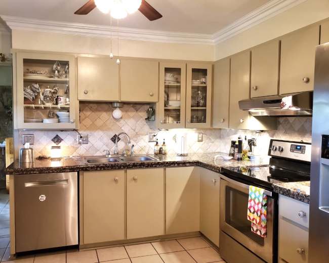 The image shows a kitchen with beige cabinets, stainless steel appliances, and a tiled backsplash, featuring a sink and organized dishware in glass-fronted cabinets.