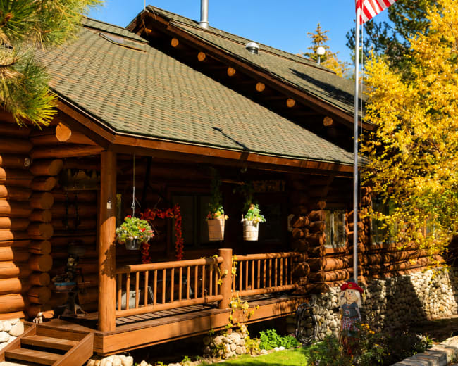 A log cabin with a flagpole displaying the American flag stands amidst autumn foliage and a well-kept garden.