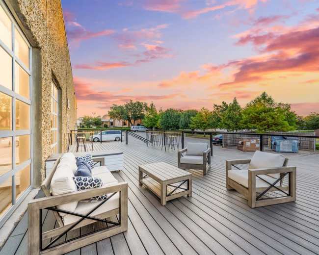 A deck with modern wooden furniture overlooks a colorful sunset sky with orange and pink hues.