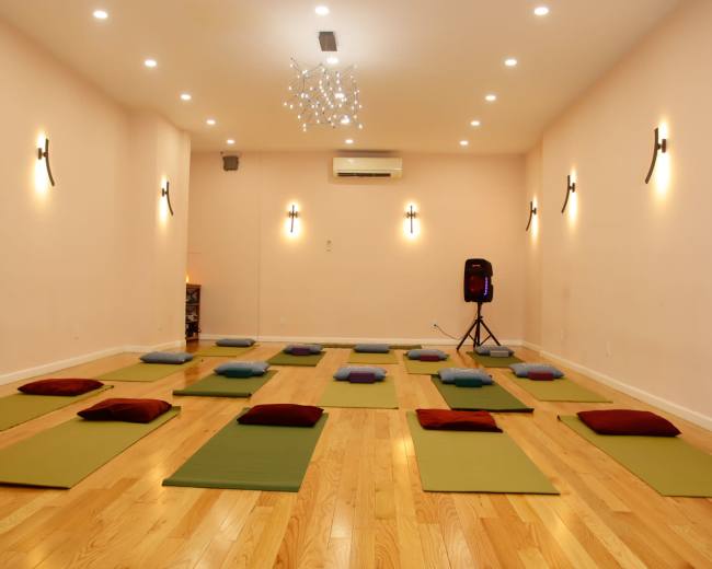 The image shows a spacious yoga studio with wooden floors, arranged mats, and cushions set up for a class.