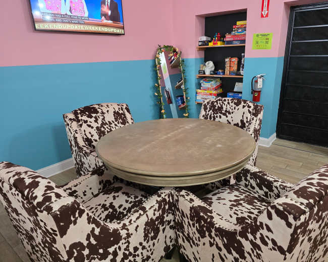 The image shows a round table surrounded by four cow-patterned chairs in a room with pink and blue walls, a television, and a shelf filled with games.