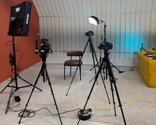 The image shows a studio setup with three cameras on tripods, a chair in the center, and various lighting equipment against a backdrop of red and white walls.