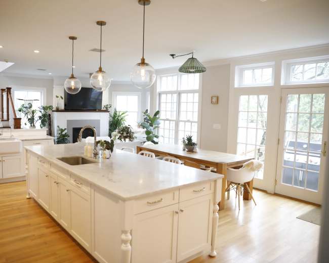 The image shows a bright, modern kitchen with a large island, marble countertops, pendant lighting, and an adjacent dining area with a wooden table and chairs.