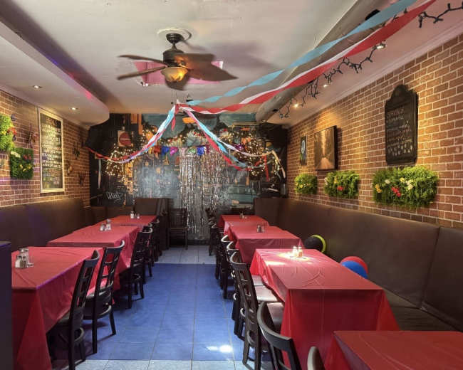 A narrow restaurant space features long tables covered with red tablecloths, surrounded by brick walls adorned with green plants and festive decorations.