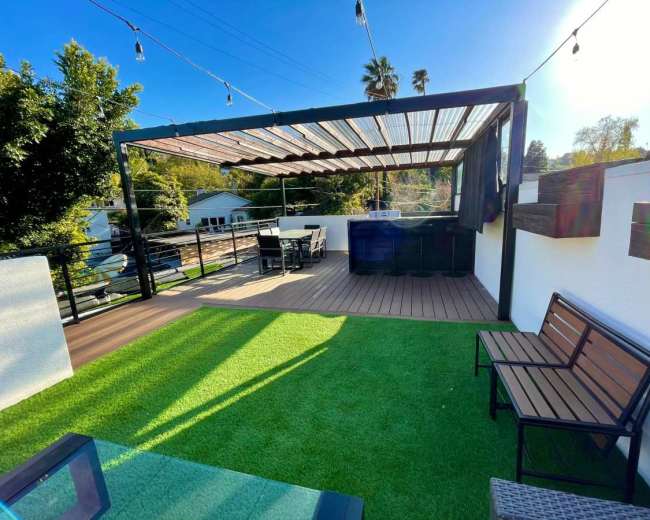 The image shows a rooftop deck with artificial grass, a shaded seating area, and a dining setup.