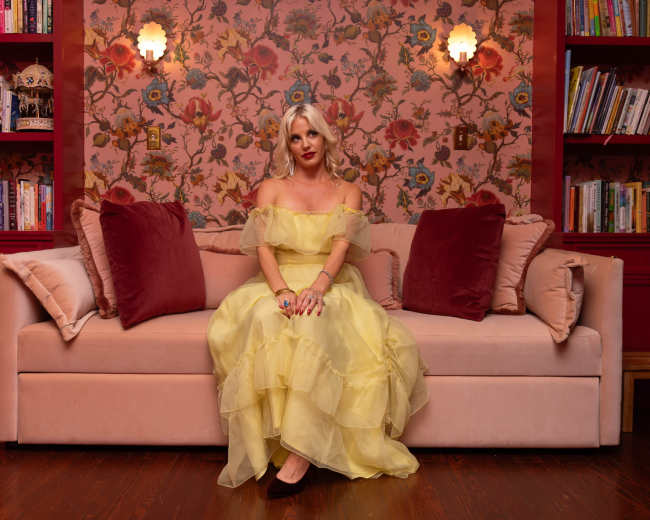 A woman in a yellow dress sits on a pink sofa in a room adorned with floral wallpaper and bookshelves.