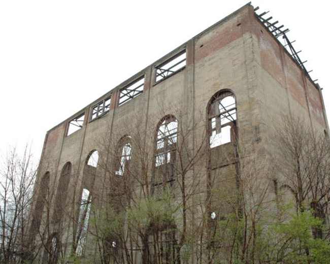 The remains of a tall, abandoned industrial building with partially collapsed walls and overgrown vegetation surrounding it.