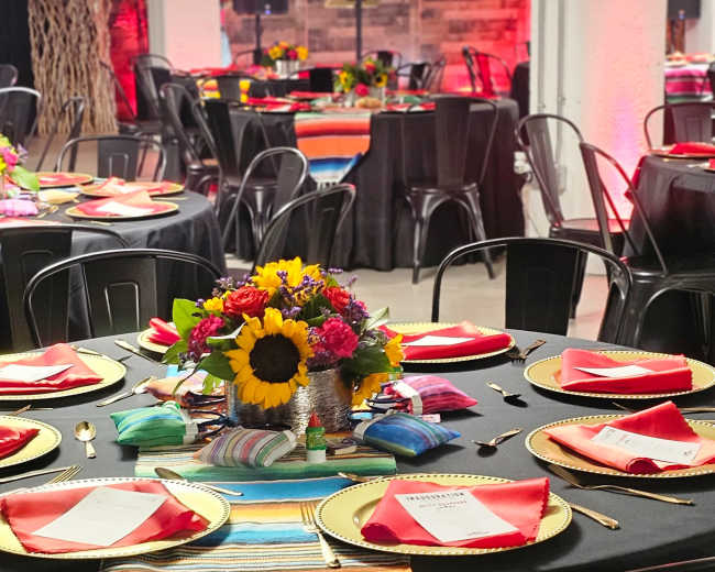 A banquet hall is set up with multiple round tables adorned with colorful tablecloths, gold chargers, and floral centerpieces, illuminated by soft red lighting.
