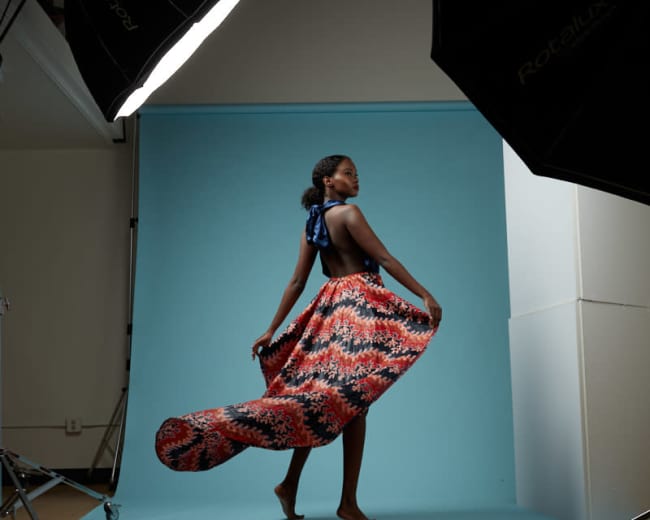 A model poses gracefully in a colorful flowing dress against a light blue backdrop, surrounded by studio lighting equipment.