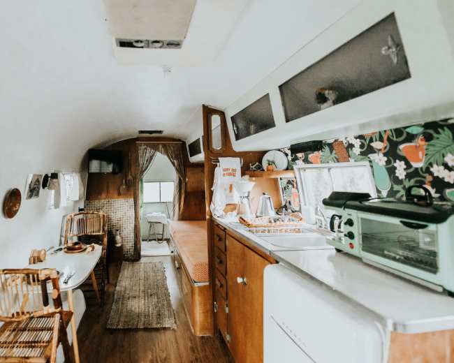 The interior of a vintage trailer with wooden cabinetry, a small dining table, and a kitchenette featuring floral wallpaper.