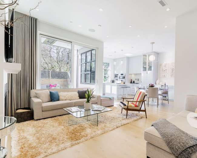A modern living room features a light gray sofa and armchair, a glass coffee table, and large windows that overlook a well-lit kitchen area.