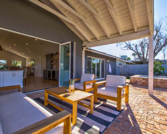 A spacious outdoor seating area features wooden furniture and opens up to an interior kitchen and living space with large windows and a bright atmosphere.