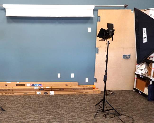 A photography studio setup with two softbox lights on stands positioned in front of a plain blue wall, and cardboard boxes placed on the floor.