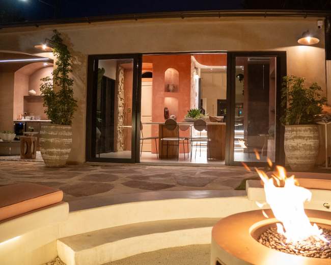 A modern outdoor space features a fire pit in the foreground with flames, and a view of a warmly-lit interior room visible through sliding glass doors.